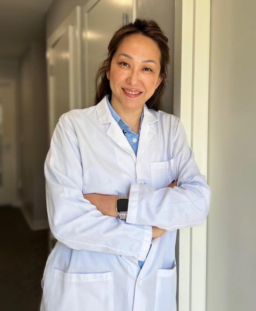 Dr. Yusun Gohard, DDS wearing a white lab coat and arms crossed, leaning against a doorway in an office corridor.
