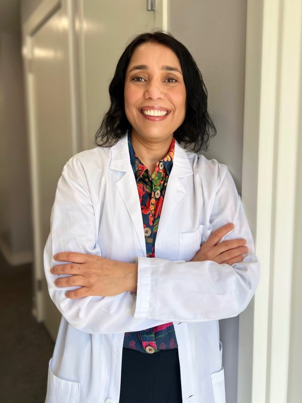 Dr. Devika Bhola wearing a white lab coat over a patterned top, smiling with arms crossed in an office setting.