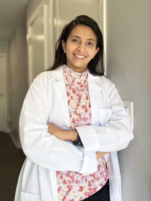 Dr. Shah wearing a white lab coat over a floral blouse, smiling with arms crossed in a hallway.