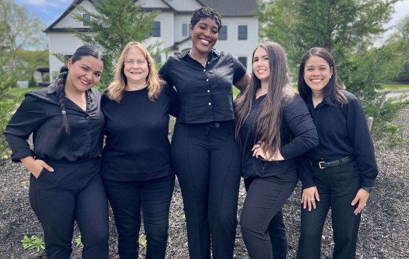 Five female front desk members smiling and posing together outdoors in matching black uniforms.